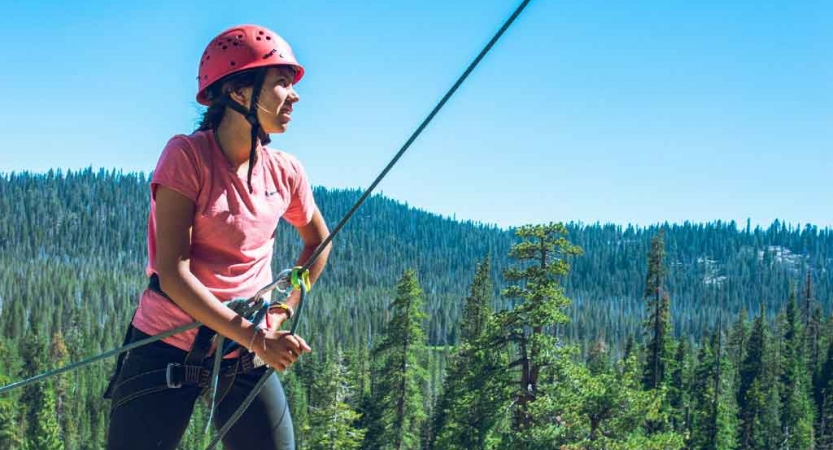 A person wearing safety gear is secured by ropes while belaying an unseen climber. There is an evergreen forest in the background. 